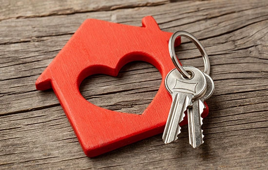 Red house-shaped keychain with heart cutout and two silver keys attached, placed on weathered wooden surface